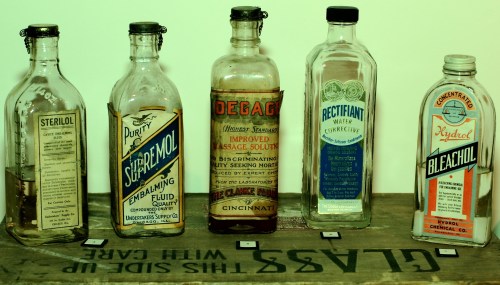 A row of vintage glass bottles labeled with various chemical and medicinal names on a wooden surface.