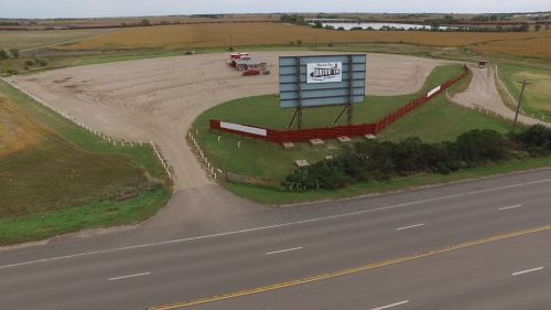 Aerial view of a drive-in theater with a large screen, surrounded by open fields and a road in the foreground.