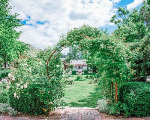 A lush garden with a green archway, leading to a scenic view of a house and blue sky with fluffy clouds.