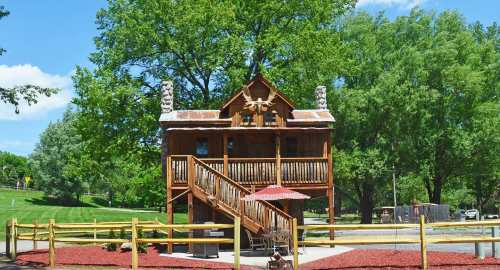 A rustic wooden cabin with a metal roof, surrounded by trees and a fenced area, featuring a staircase and picnic table.