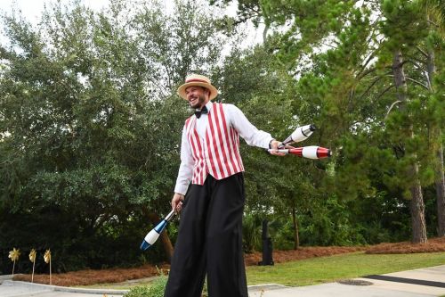 A performer in a striped vest and bow tie juggles colorful clubs outdoors, surrounded by trees.