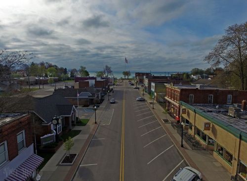 A quiet small town street lined with shops, trees, and a view of the lake under a cloudy sky.