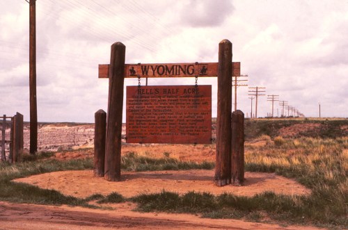 Sign for Hell's Half Acre in Wyoming, featuring wooden posts and a scenic landscape with power lines in the background.