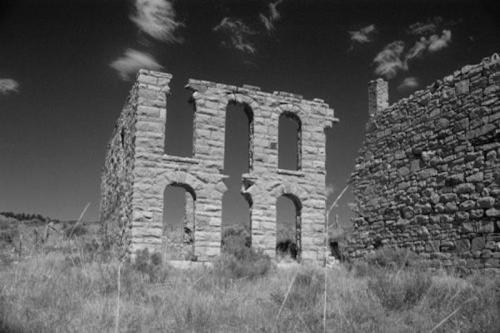 Black and white image of stone ruins with arched windows, set against a cloudy sky and grassy landscape.