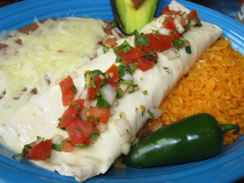 A blue plate with a cheese-covered burrito topped with diced tomatoes and cilantro, served with rice, beans, and a jalapeño.