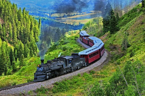 A vintage steam train winds through lush green hills, surrounded by trees and mountains, with smoke billowing from the engine.