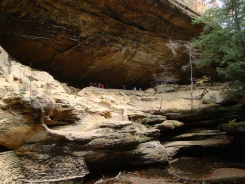 A rocky cliff with a large overhang, surrounded by trees and a small stream, with people exploring the area above.