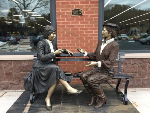 Two bronze statues of a man and woman sitting on a bench, engaged in conversation, set against a brick wall.
