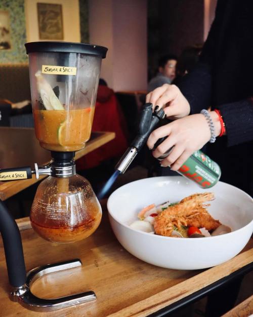 A person uses a torch on a blender while preparing a dish with shrimp and vegetables in a bowl.