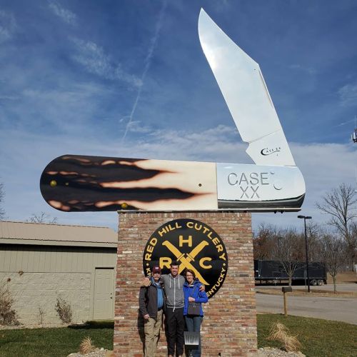 A couple stands in front of a large knife sculpture at Red Hill Cutlery, with a blue sky in the background.