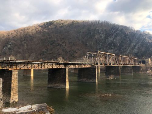 A rusty metal bridge spans a river, with a rocky hillside and cloudy sky in the background.