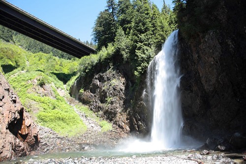 A waterfall cascades down rocky cliffs, surrounded by lush greenery, with a highway bridge overhead.
