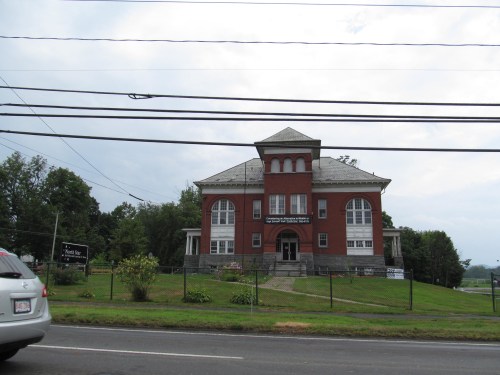 Historic red brick building with large windows, surrounded by greenery and a fence, under a cloudy sky.