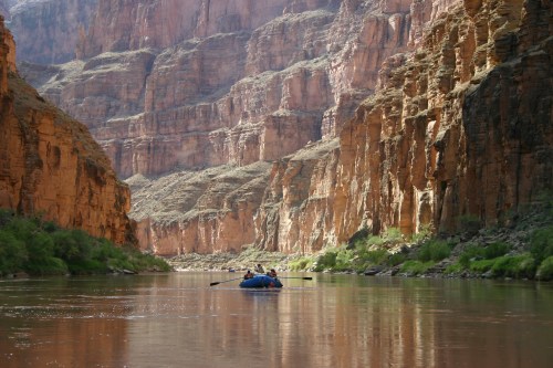 A blue raft floats on a calm river surrounded by towering red rock cliffs and lush greenery.