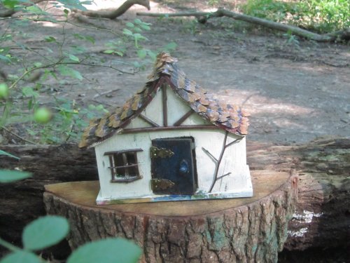 A small, whimsical wooden house with a shingled roof, placed on a tree stump in a forest setting.