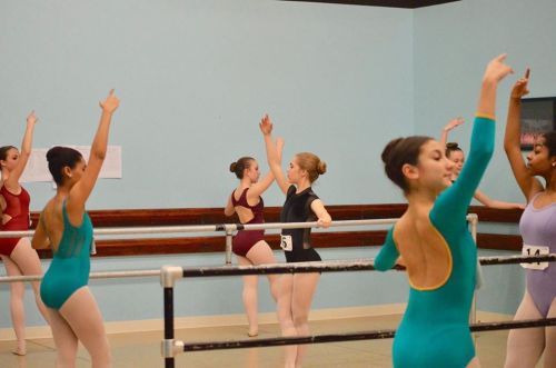 A group of young ballet dancers practice at the barre in a studio, wearing colorful leotards and tights.