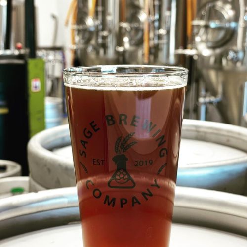 A glass of amber beer with the logo of Sage Brewing Company, set against brewing equipment in the background.