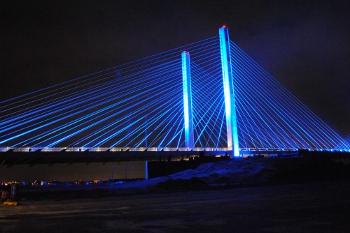 A brightly lit suspension bridge at night, featuring blue lights illuminating its cables and towers against a dark sky.