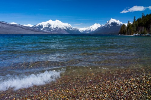 A serene lake with colorful pebbles in the foreground, surrounded by snow-capped mountains under a clear blue sky.