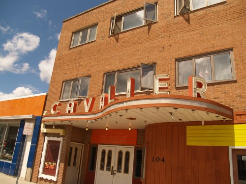 Exterior of the Cavalier Theater, featuring a marquee and large windows, against a blue sky with clouds.