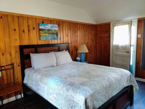 Cozy bedroom with wooden walls, a double bed, a lamp, a chair, and a door leading outside.