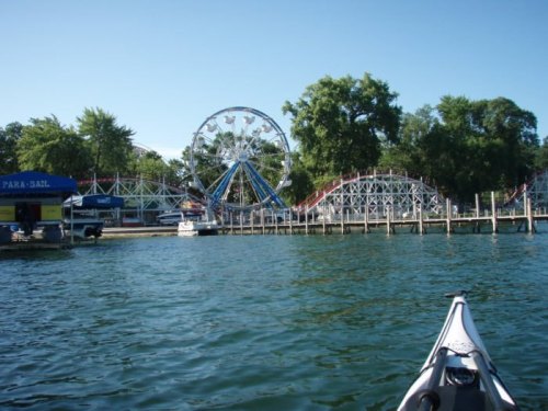 A view from a kayak of a lakeside amusement park featuring a ferris wheel and roller coaster under a clear blue sky.