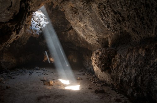 Sunlight streams through a cave opening, illuminating rocky walls and a sandy floor.