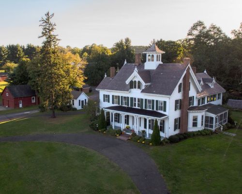 Aerial view of a large white Victorian house with a wraparound porch, surrounded by green lawns and trees.