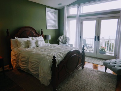 Cozy bedroom with a green wall, a wooden bed, and large windows overlooking a balcony. Soft bedding and a chair add comfort.