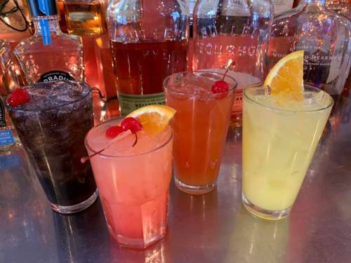 Four colorful cocktails with garnishes, set against a backdrop of various liquor bottles.