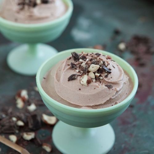Two green dessert cups filled with chocolate mousse, topped with chopped nuts and chocolate shavings on a textured surface.