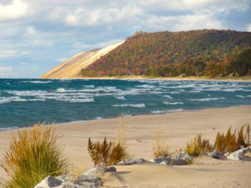 A sandy beach with gentle waves, framed by colorful autumn trees and a large dune in the background under a cloudy sky.