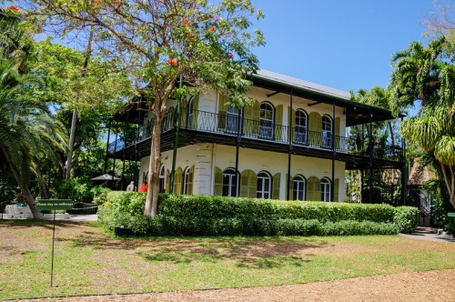 A two-story house surrounded by lush greenery and trees, featuring balconies and a clear blue sky.