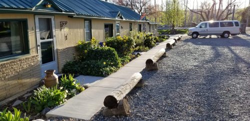 A pathway lined with logs leads to a row of cabins, surrounded by greenery and a parked van nearby.