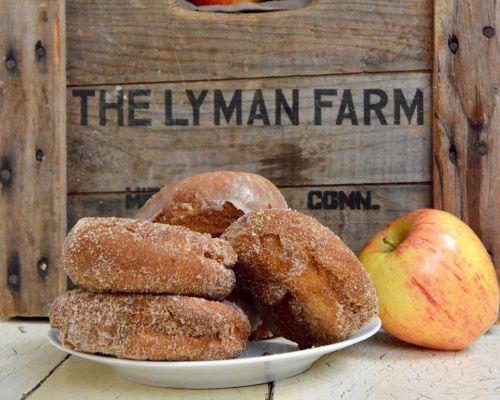 A plate of cinnamon-sugar donuts next to a fresh apple, with a wooden crate in the background labeled "The Lyman Farm."