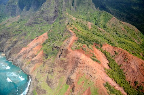 Aerial view of lush green mountains and rugged cliffs meeting the ocean, with waves crashing against the shore.