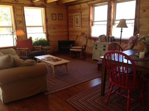 Cozy wooden living room with a sofa, rocking chairs, a table, and large windows letting in natural light.