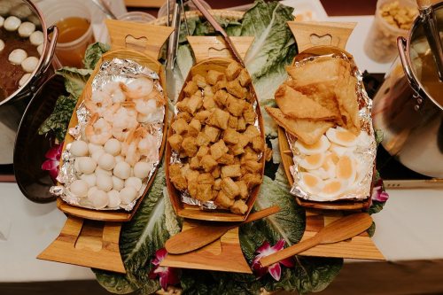 Three wooden trays filled with shrimp, fried tofu, and crispy snacks, surrounded by leafy greens and garnished with flowers.