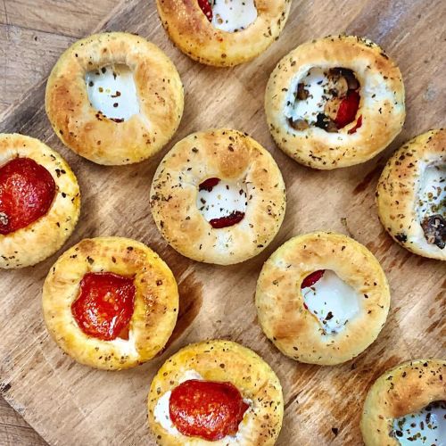 A wooden board with various mini pastry cups filled with cheese, pepperoni, and herbs.