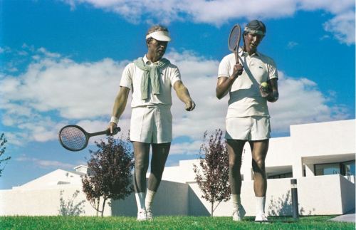 Two men in tennis attire walking on a grassy court, holding rackets, with a modern building and blue sky in the background.