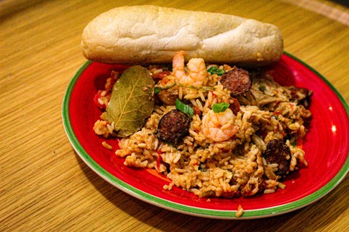 A vibrant red plate filled with shrimp and sausage rice, garnished with bay leaves, alongside a piece of bread.