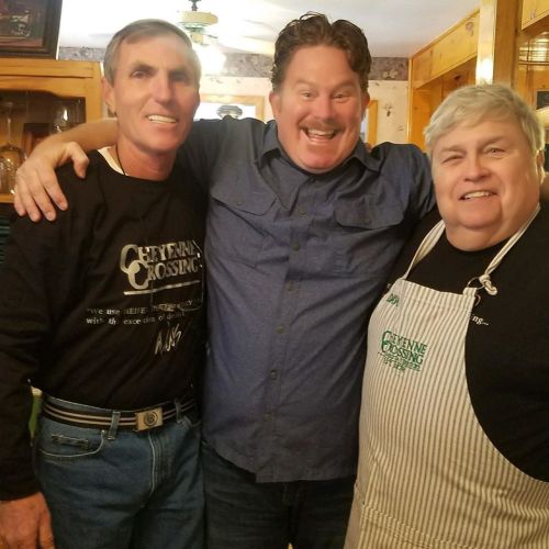 Three smiling individuals pose together in a cozy kitchen setting, with one wearing an apron and the others in casual attire.