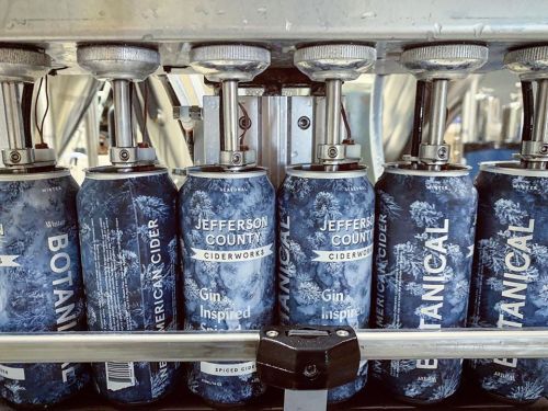 Cans of Jefferson County Botanical Cider on a production line, featuring a blue design and labeled "Gin Inspired."