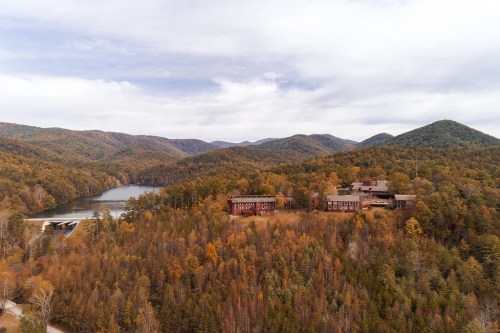 Aerial view of a serene lake surrounded by autumn-colored mountains and a rustic lodge nestled among trees.
