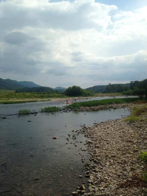 A serene river scene with rocky banks, lush greenery, and distant mountains under a cloudy sky.