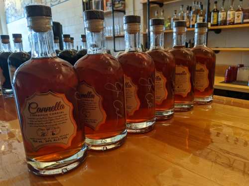 A row of bottles labeled "Carmella Cinnamon Cordial" on a wooden bar, showcasing their rich amber liquid.