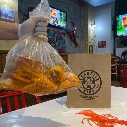 A gloved hand holds a bag of seafood, with a takeout bag from a restaurant on the table and TVs in the background.
