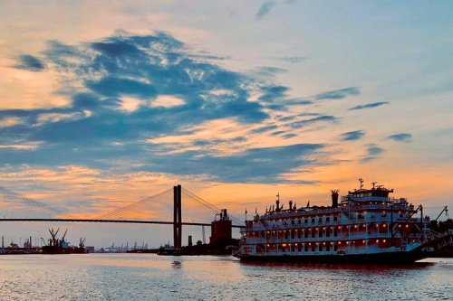 A riverboat docked near a bridge at sunset, with colorful clouds reflecting on the water.