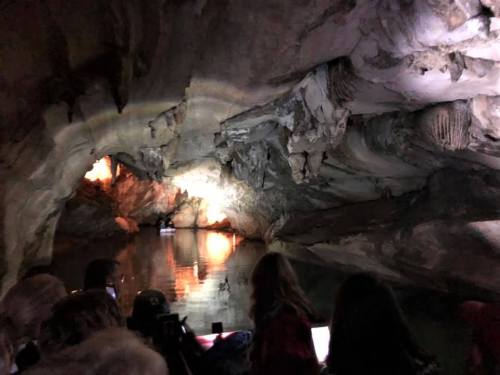 A dimly lit cave with water reflecting light, viewed from a boat with people in the foreground.
