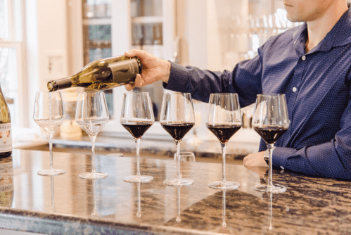 A person pours wine from a bottle into a row of six glasses on a countertop, showcasing different wine types.